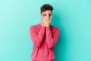 Mavi arka planda izole edilmiş genç beyaz adam kameraya parmaklarıyla göz kırpıyor, yüzünü kapatmaktan utanıyor..