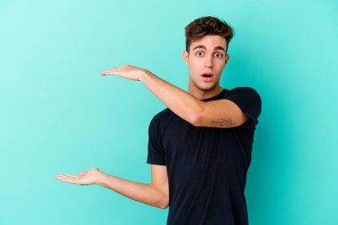 Mavi arka planda izole genç beyaz adam şok ve hayret içinde elleri arasında bir kopya boşluk tutuyor.