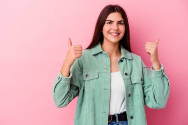 Pembe arka planda izole edilmiş genç beyaz kadın gülümsüyor ve başparmağını kaldırıyor.