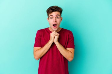 Kafkasyalı genç adam mavi arka planda izole edilmiş şans için dua ediyor, şaşırmış ve ağzını açıp önüne bakıyor..
