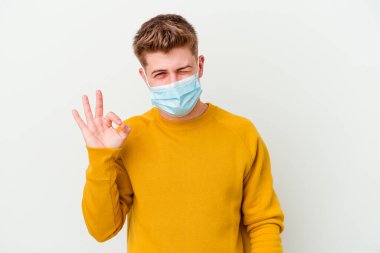 Koronavirüs için maske takan genç bir adam beyaz arka planda izole edilmiş bir şekilde bir göz kırpıyor ve elleriyle iyi bir jest yapıyor..