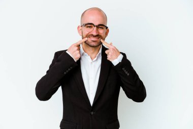 Genç, beyaz, kel bir adam mavi arka planda izole edilmiş gülümsüyor ve parmaklarını ağzına doğrultuyor..