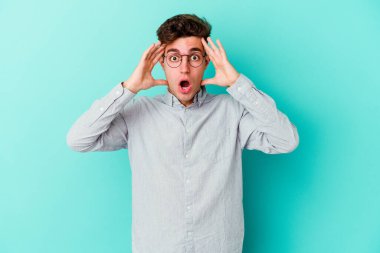 Mavi arka planda izole edilmiş genç beyaz adam yüzünün yarısını palmiye ile kaplayarak eğleniyor..