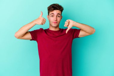 Kafkasyalı genç adam mavi arka planda izole edilmiş, baş parmağını kaldırıyor, zor seçim konsepti.