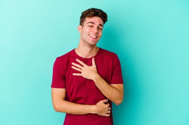 Mavi arka planda izole edilmiş genç beyaz adam mutlu bir şekilde gülüyor ve ellerini karnının üstünde tutarak eğleniyor..