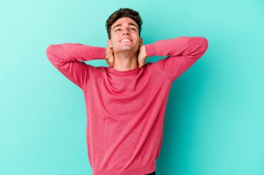 Kafkasyalı genç adam mavi arka planda izole edilmiş. Kendine güveniyor, elleri başının arkasında..