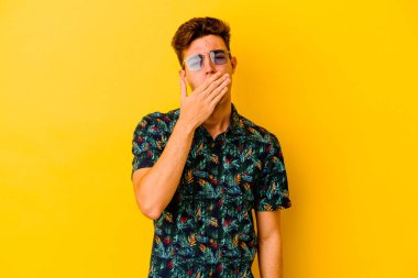 Sarı arka planda izole edilmiş Hawaii gömleği giyen genç beyaz bir adam esneyerek ağzını eliyle kapatan yorgun bir jest gösteriyor..