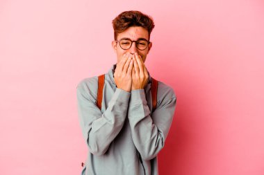 Pembe arka planda izole olmuş genç bir öğrenci bir şeye gülüyor, elleriyle ağzını kapatıyor..