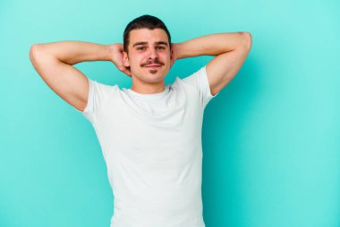 Kafkasyalı genç adam mavi arka planda izole edilmiş. Kendine güveniyor, elleri başının arkasında..