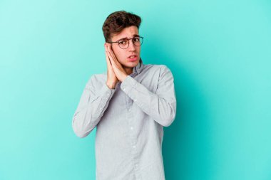 Mavi arka planda izole edilmiş genç beyaz adam esniyor. Ağzı eliyle kapatan yorgun bir hareket sergiliyor..