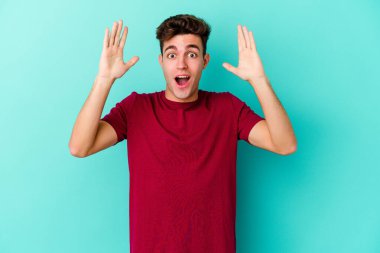 Mavi arka planda izole edilmiş genç beyaz bir adam zaferi ya da başarıyı kutluyor, şaşırıyor ve şok oluyor..