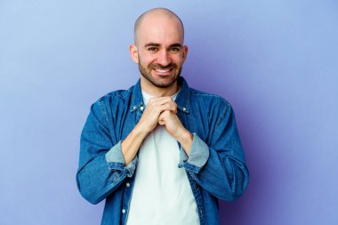 Mor arka planda izole genç beyaz kel adam ellerini çenesinin altında tutuyor, mutlu bir şekilde yana bakıyor..