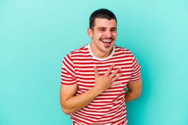 Mavi arka planda izole olmuş genç beyaz adam yüksek sesle gülüyor ve elini göğsüne koyuyor..