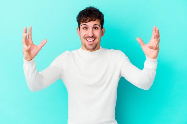 Mavi arka planda izole edilmiş genç beyaz adam hoş bir sürpriz alıyor, heyecanlı ve el kaldırıyor..