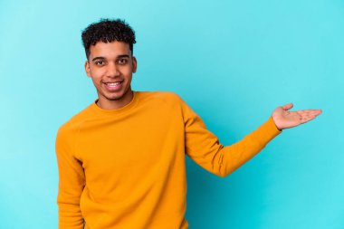Genç Afro-Amerikalı kıvırcık adam mavi üzerine izole edilmiş bir şekilde bir avuç içinde fotokopi alanı gösteriyor ve başka bir elini belinde tutuyor..