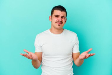 Kafkasyalı genç adam mavi arka planda izole edilmiş sorgulama jestinde şüphe ediyor ve omuz silkiyor..