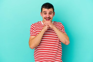 Mavi arka planda izole edilmiş genç beyaz adam ellerini çenesinin altında tutuyor, mutlu bir şekilde yana bakıyor..