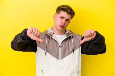 Sarı arka planda izole edilmiş genç beyaz adam baş parmağını aşağı gösteriyor ve hoşlanmadığını ifade ediyor..