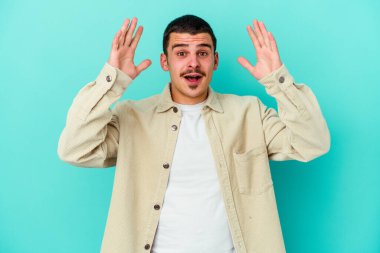 Mavi arka planda izole edilmiş genç beyaz bir adam zaferi ya da başarıyı kutluyor, şaşırıyor ve şok oluyor..