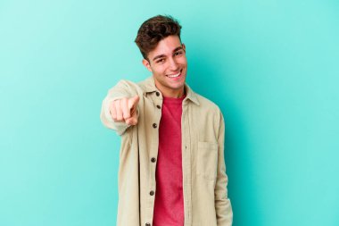 Mavi arka planda izole edilmiş genç beyaz adam parmaklarıyla ön tarafı işaret ediyor..
