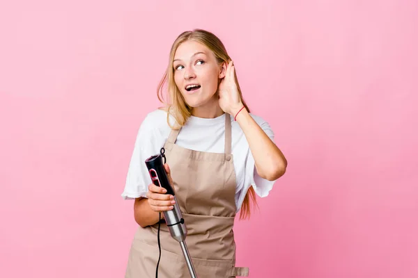 Young russian cook woman holding an electric mixer isolated trying to listening a gossip.