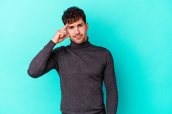 Молодой кавказский человек изолирован на синем фоне, указывая пальцем на храм, думая, сосредоточен на задаче.