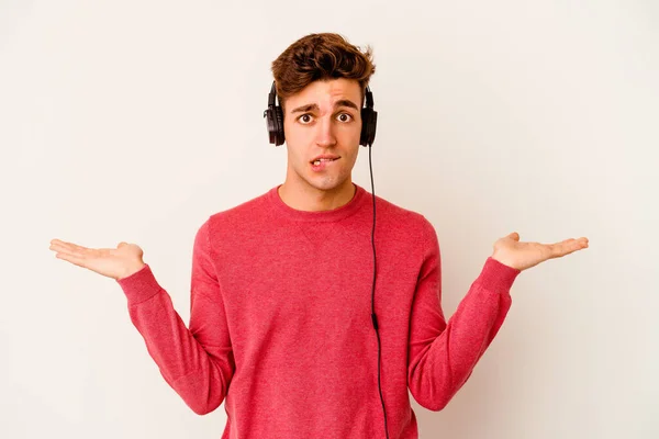 Jovem Caucasiano Ouvindo Música Isolada Fundo Branco Confuso Duvidoso Ombros — Fotografia de Stock