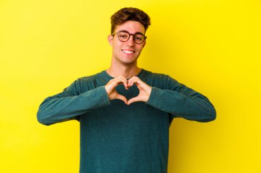 Sarı arka planda izole edilmiş genç beyaz adam gülümsüyor ve elleriyle kalp şeklini gösteriyor..