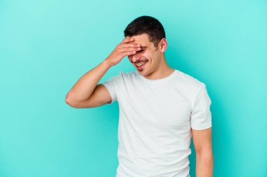 Mavi arka planda izole edilmiş genç beyaz adam kameraya parmaklarıyla göz kırpıyor, yüzünü kapatmaktan utanıyor..