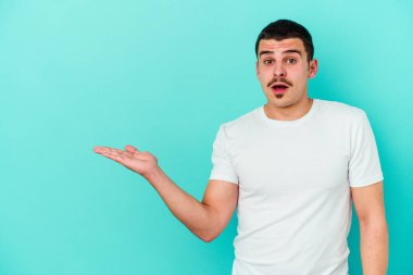 Mavi arka planda izole edilmiş genç beyaz adam, avuç içi fotokopi alanını elinde tutmaktan etkilenmiş..