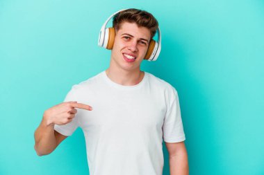 Mavi arka planda kulaklıklı müzik dinleyen beyaz bir adam gömlek fotokopi alanını işaret ediyor, gururlu ve kendinden emin.