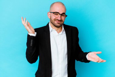 Genç, beyaz, kel bir adam mavi arka planda izole edilmiş. Kafası karışmış ve omuz silkiyor..