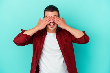 Mavi arka planda soyutlanmış genç beyaz bir adam elleriyle gözlerini kapatıyor, geniş bir gülümsemeyle bir sürpriz bekliyor..