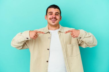 Mavi arka planda izole edilmiş genç beyaz adam parmağını göstererek şaşırdı, geniş bir gülümsemeyle..