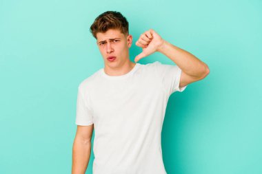 Mavi arka planda izole edilmiş genç beyaz bir adam baş parmağını aşağı gösteriyor ve hoşlanmadığını ifade ediyor..