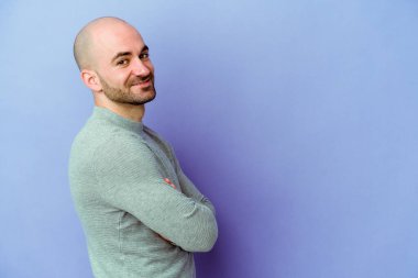 Mor arka planda soyutlanmış beyaz kel bir adam kollarını kavuşturarak gülümsüyor..