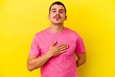 Beyaz tenli genç adam sarı arka planda izole edilmiş yemin ediyor, elini göğsüne koyuyor..