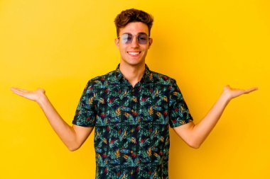 Sarı arka planda izole edilmiş Hawaii tişörtü giyen genç beyaz bir adam kollarıyla terazi yapıyor, mutlu ve kendinden emin hissediyor..