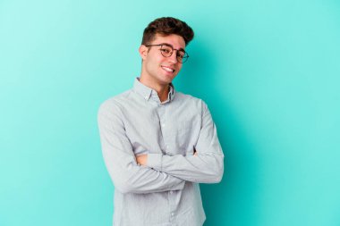 Mavi arka planda kendini güvende hisseden, kararlılıkla kollarını kavuşturan genç beyaz bir adam..