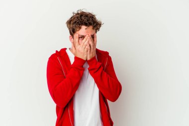 Beyaz arka planda izole edilmiş genç beyaz bir adam parmaklarının arasından korkmuş ve gergin bir şekilde göz kırpıyor..