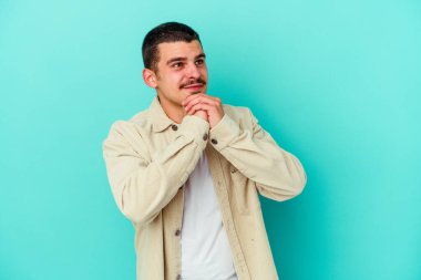 Mavi arka planda izole edilmiş genç beyaz adam ellerini çenesinin altında tutuyor, mutlu bir şekilde yana bakıyor..