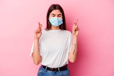 Virüs maskesi takan beyaz bir kadın şans dilemek için pembe arka planda izole edilmiş.