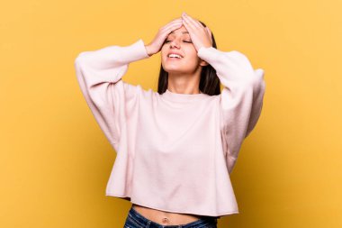 Sarı arka planda izole edilmiş genç Hintli kadın mutlu bir şekilde ellerini başının üstünde tutarak gülüyor. Mutluluk kavramı.