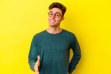 Sarı arka planda izole edilmiş genç beyaz adam selamlama jesti olarak kameraya elini uzatıyor..