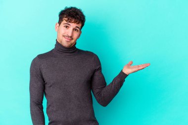 Mavi arka planda izole edilmiş genç beyaz bir adam bir palmiye üzerinde fotokopi alanı ve belinde bir el daha gösteriyor..