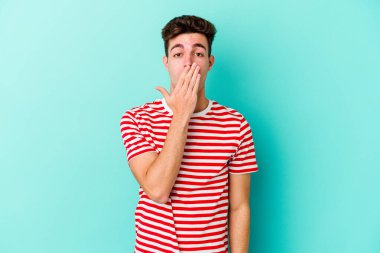 Mavi arka planda izole edilmiş genç beyaz adam şok olmuş, ağzı ellerle kaplanmış, yeni bir şey keşfetmeye hevesli..