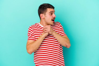Kafkasyalı genç adam mavi arka planda izole edilmiş şans için dua ediyor, şaşırmış ve ağzını açıp önüne bakıyor..