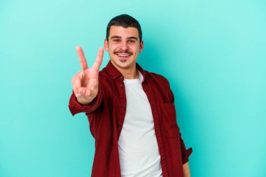 Mavi arka planda izole edilmiş genç beyaz adam zafer işareti gösteriyor ve gülümsüyor..