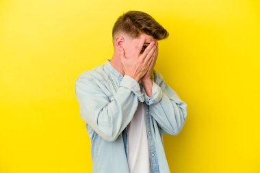 Sarı arka planda izole edilmiş genç beyaz adam parmaklarının arasından korkmuş ve gergin bir şekilde göz kırp..