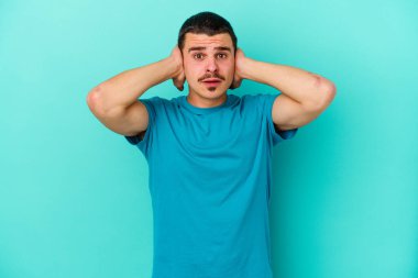 Kafkasyalı genç adam mavi arka planda izole edilmiş kulakları kulaklarıyla kapatıyor çok fazla ses duymamaya çalışıyor..